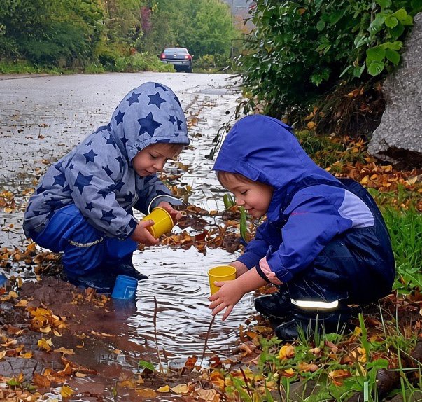 Zwei Kinder in Regenkleidung spielen mit Bechern an einer Pfütze im Herbstlaub.