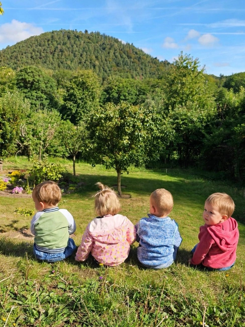 Vier Kinder sitzen nebeneinander auf der Wiese und schauen auf Apfelbaum, Wald und Berg unter blauem Himmel, das rechte Kind schaut über die Schulter zurück.
